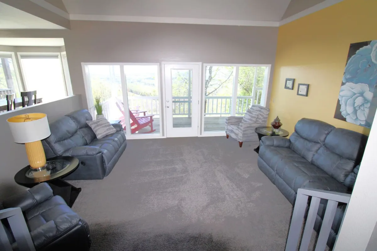 A cozy, sunlit living area with comfortable seating, vibrant wall art, a decorative lamp, and expansive windows. The view through the windows evokes the lakeside setting and enhances the inviting atmosphere for group relaxation.