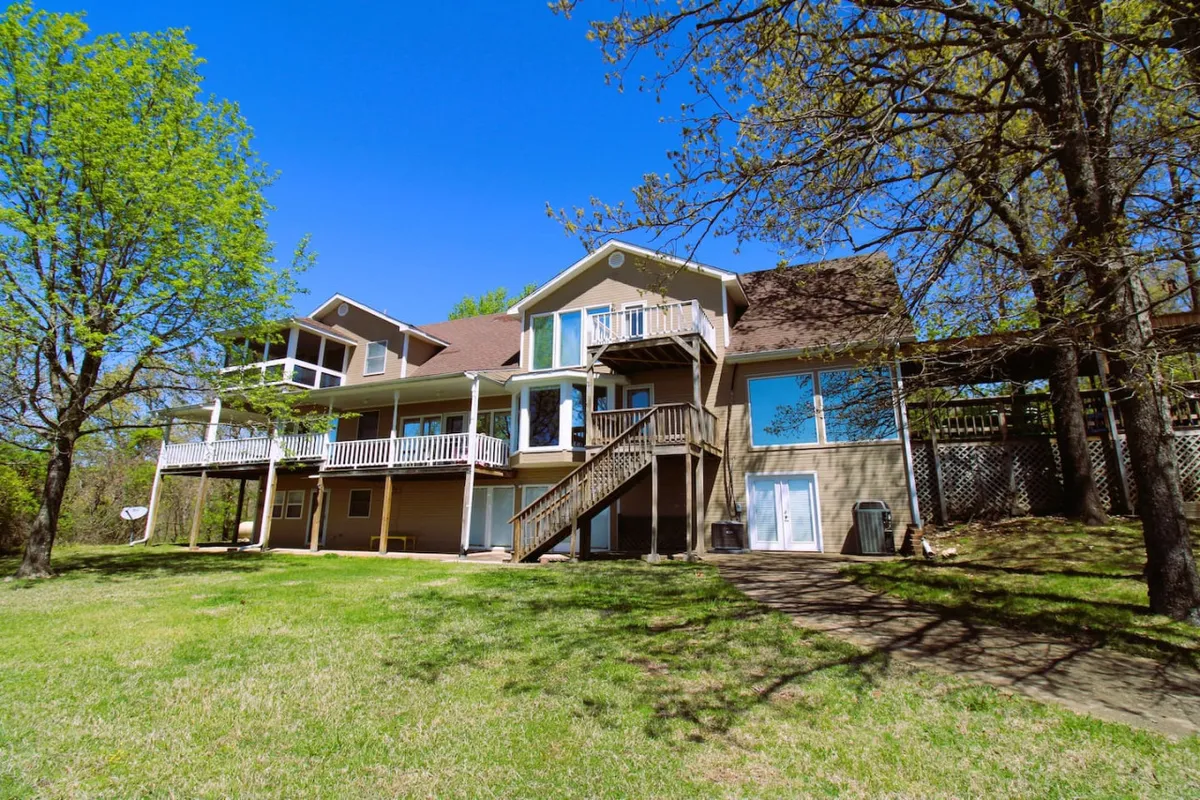 The image showcases the exterior of a spacious single-family home perched atop a gentle hill, featuring a large wrap-around deck with inviting seating areas. The lush, expansive lawn is bordered by mature trees, offering both privacy and a serene backdrop for outdoor activities. A grand staircase leads up to the deck, suggesting easy access for gatherings and relaxation. The setting is bathed in the warm glow of sunset, highlighting the tranquil atmosphere and the promise of stunning lake views.