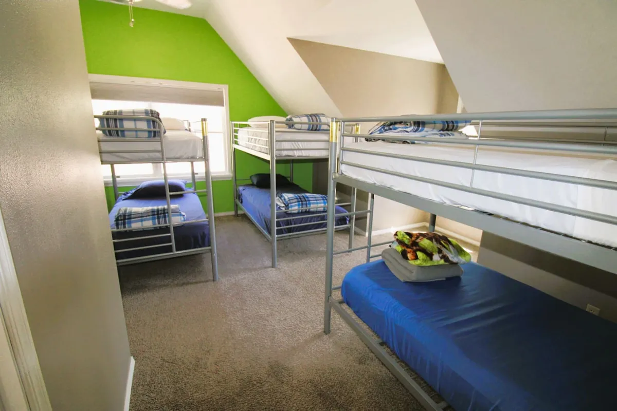 This image showcases a vibrant bunk room featuring multiple bunk beds with plaid bedding and a bright green accent wall. Natural light streams through the window, making the space feel energetic and inviting—perfect for group sleepovers or playful mornings. The room is clearly designed for flexibility and communal fun, with enough space for group activities or late-night chats.