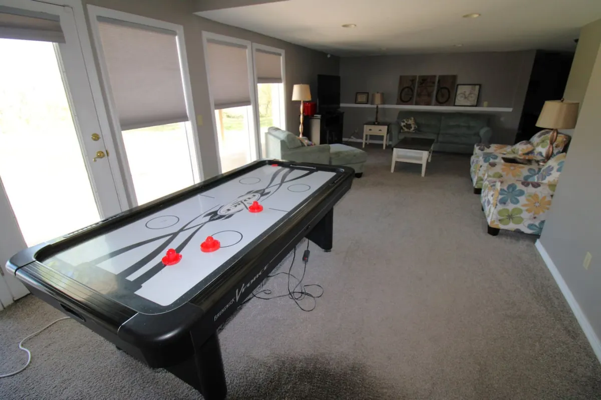 A modern air hockey table takes center stage in a bright room with a comfortable seating area and large windows that let in plenty of natural light. The scene evokes an energetic, playful mood perfect for gatherings and tournaments.