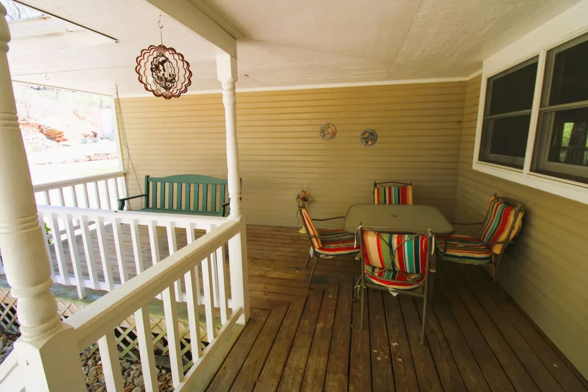 A cozy porch area features colorful striped chairs, a wooden table, a swing bench, and decorative wall plates. The setup suggests a perfect spot for socializing, sharing meals, or simply relaxing while enjoying the fresh air and views.