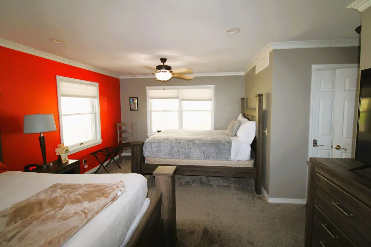 A stylish bedroom featuring a bed with gray bedding, a bright orange accent wall, and plenty of natural light streaming through large windows. The wooden furniture adds warmth, while the overall ambiance feels relaxed and inviting—appealing for adults or couples seeking comfort and privacy.