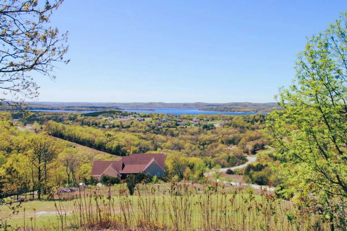 A sweeping view of Table Rock Lake with rolling hills, a house nestled among trees, and the serene water reflecting the expansive sky. This vantage point puts the lake and the natural beauty of the area center stage, inviting guests to soak in scenic views from the property.